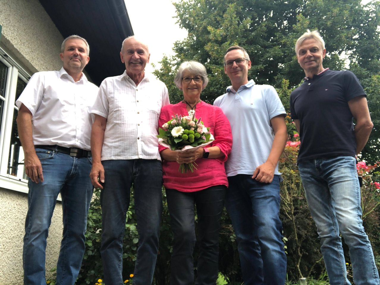 Fünf Menschen stehen vor einem Haus mit Garten und lächeln. Eine Frau in der Mitte hält einen Blumenstrauß. Die Männer tragen weiße und blaue Polo-Shirts.