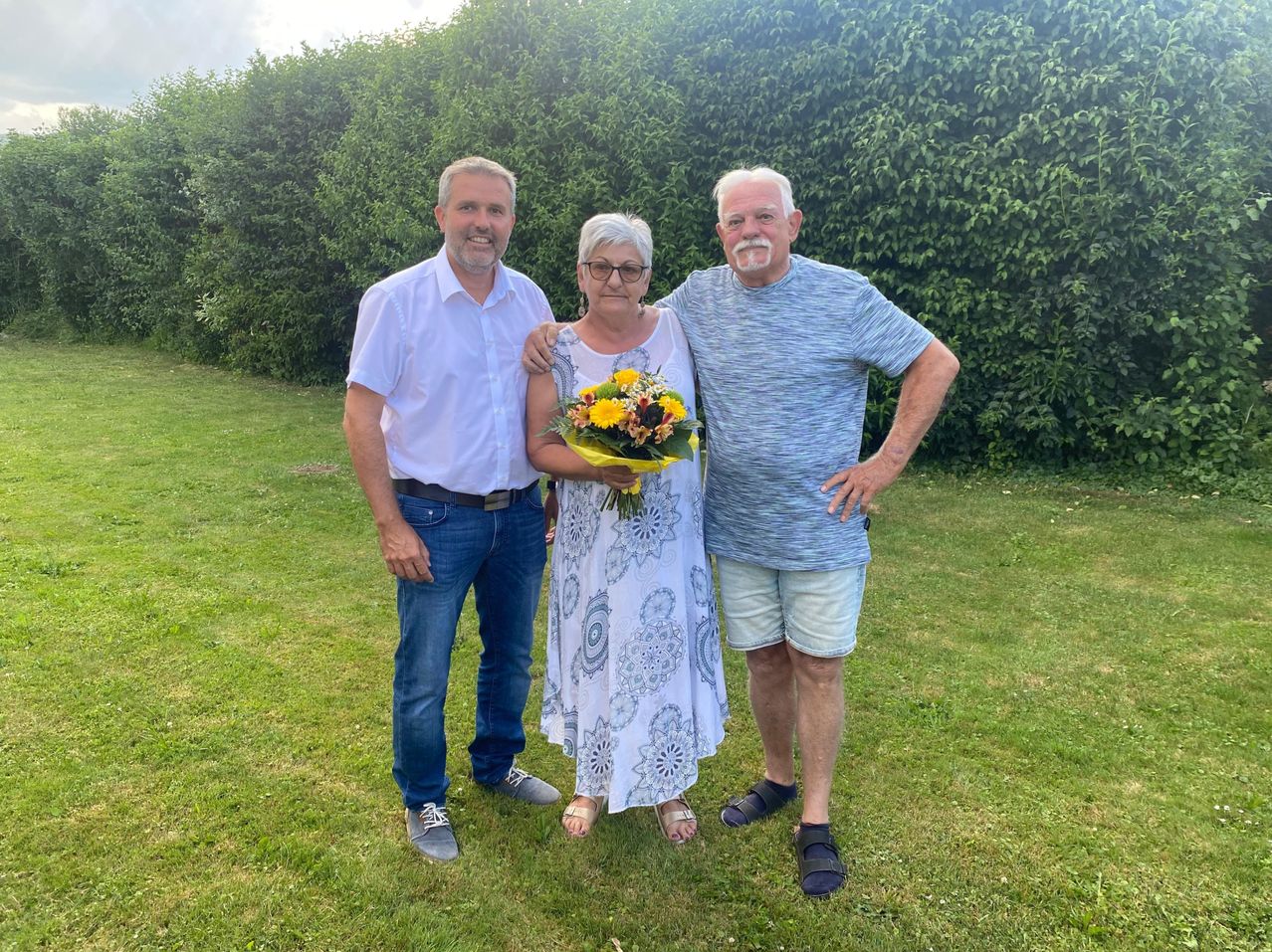 Drei Menschen stehen lächelnd in einem Grasbereich, mit einem Busch im Hintergrund. Eine Frau hält einen Blumenstrauß.