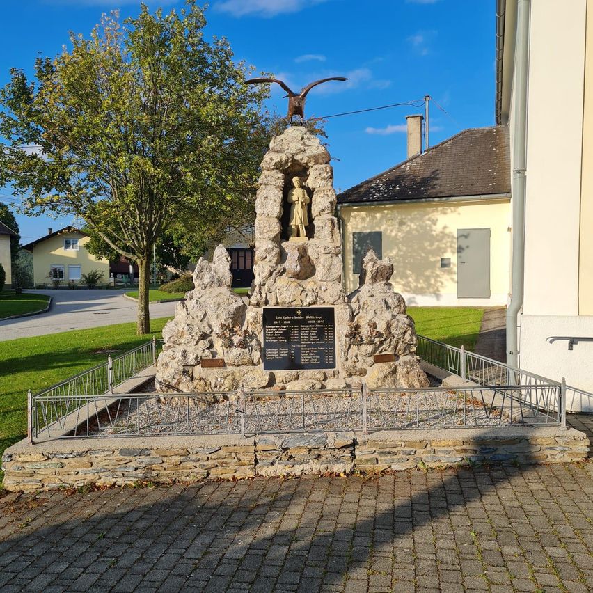 A monument with a statue at its center, surrounded by rocks and a metal fence. A tree is in the background. The sky is blue with some clouds.