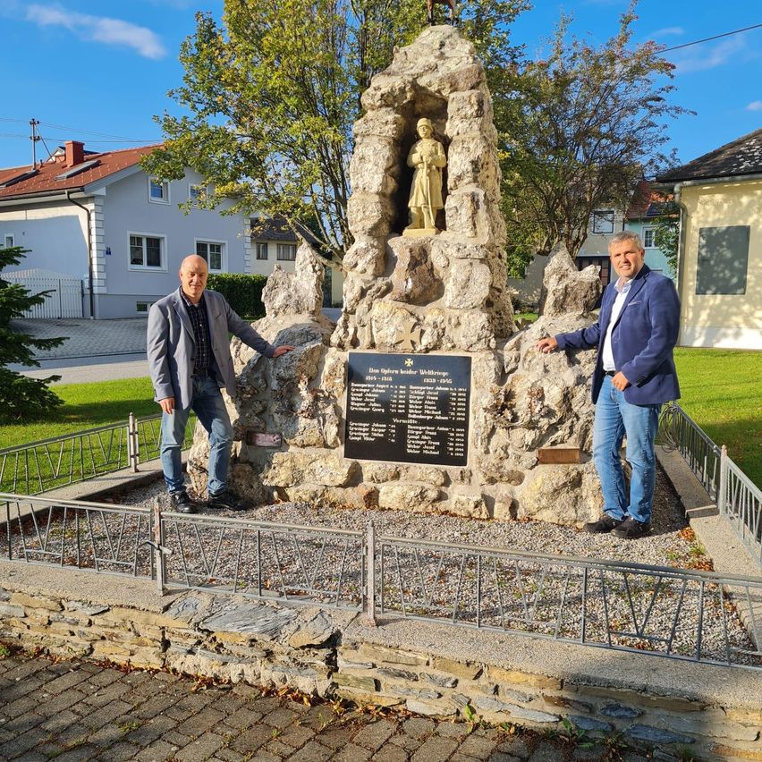 Two men stand before a monument in a park, both smiling. The monument is made of stone and has a statue of a soldier. Behind them, there are houses and trees.
