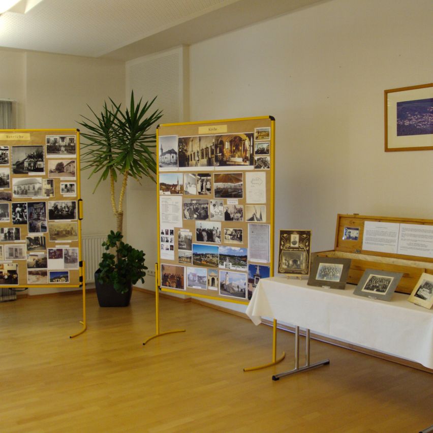 Ein Raum zeigt zwei gelbe Tafeln mit vielen Fotos, neben einer Topfpflanze. Ein Tisch mit einer weißen Tischdecke trägt einige gerahmte Fotos. Ein Holzkasten steht auf dem Tisch.