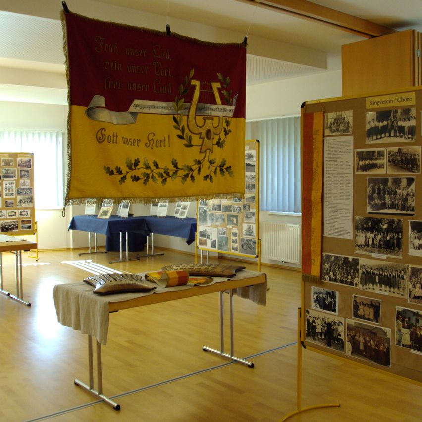 Ein Raum mit einem Tisch, der mit Tüchern und Gegenständen bedeckt ist, einem rot-gelben Banner und einem Aushang mit Bildern. Der Raum hat Holzböden und weiße Wände.