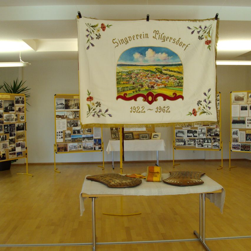 Ein Ausstellungssaal mit einem Banner, das ein Dorfbild und die Jahre 1922-1962 zeigt. Der Raum hat Holzböden und weiße Wände. Auf beiden Seiten des Raumes befinden sich Fotodisplays. In der Mitte des Raumes steht ein Tisch mit Gegenständen darauf.