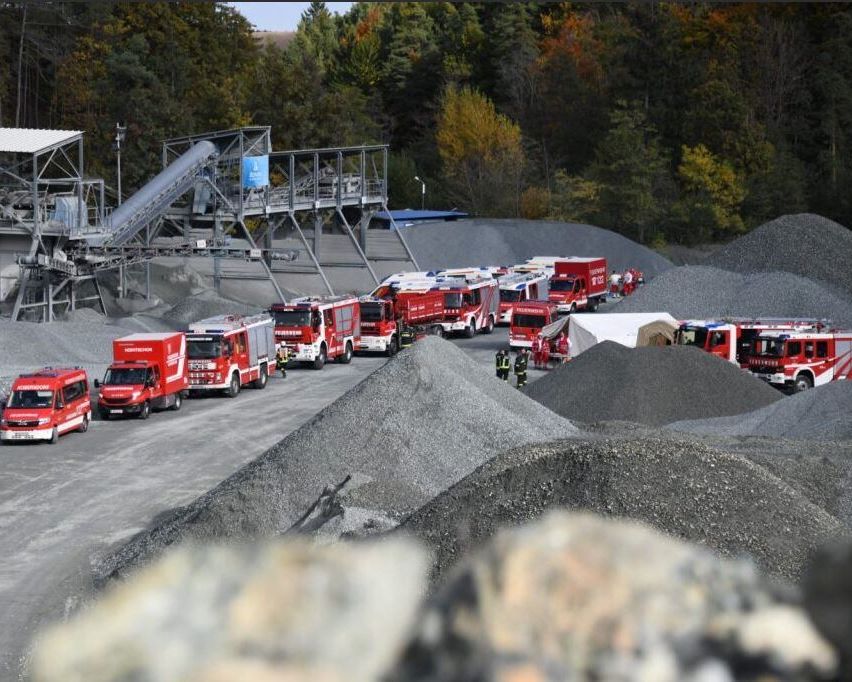 Eine Reihe roter Feuerwehrfahrzeuge, die in einem Kieswerk geparkt sind, mit Notfallpersonal in der Nähe, umgeben von einem Wald und Hügeln im Hintergrund.
