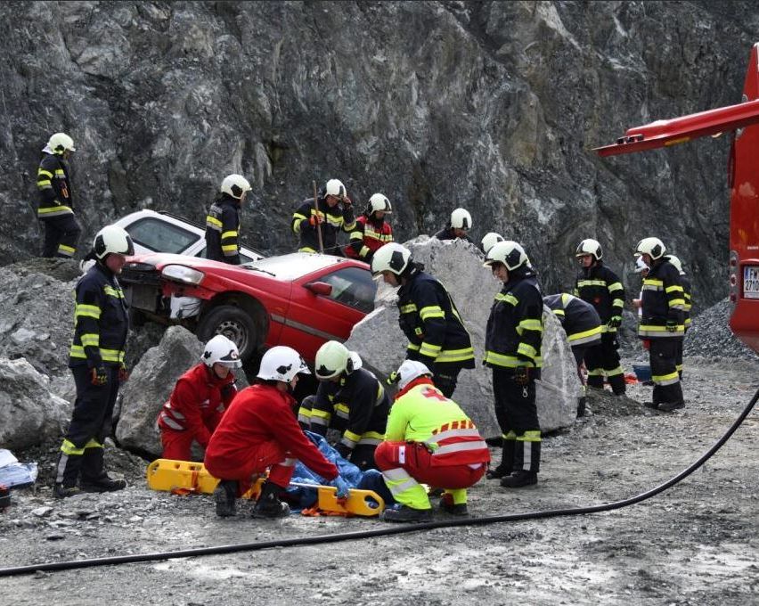 Einsatzkräfte in Helmen und Uniformen kümmern sich um einen Unfall mit einem roten Auto, mit Felsen und Steinen in der Nähe.