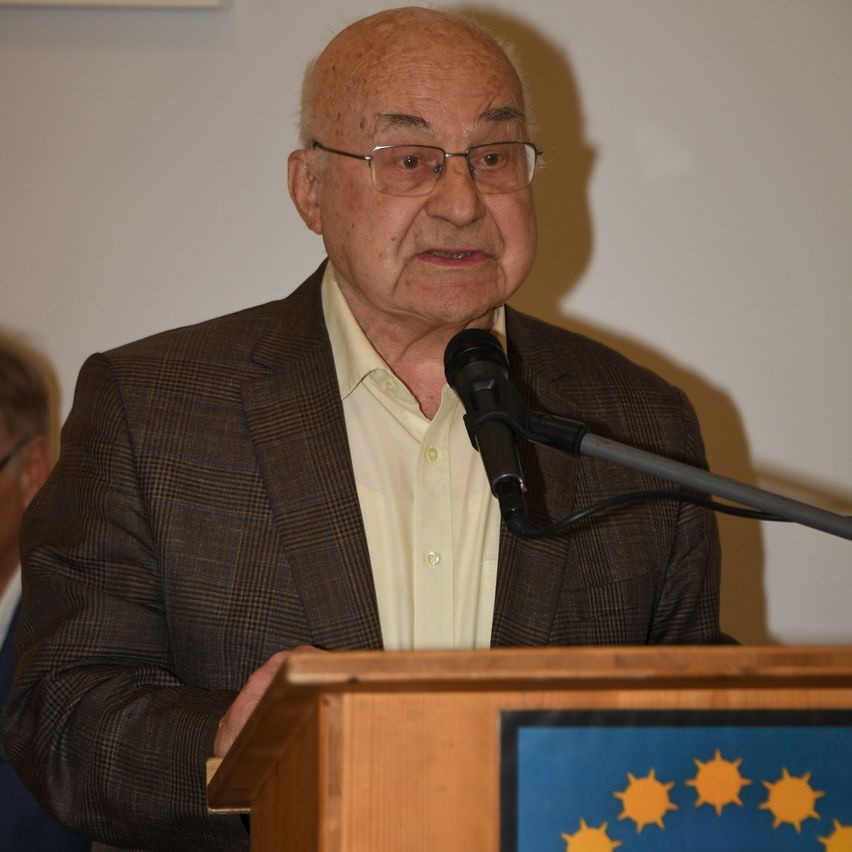 Ein älterer Mann mit Brille und einer braunen Jacke spricht an einem Podium mit Mikrofon, EU-Flagge im Hintergrund.