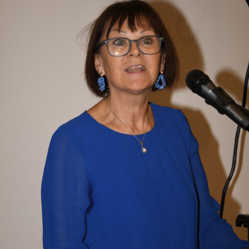 Eine ältere Frau mit kurzen braunen Haaren steht vor einem Mikrofon und trägt eine blaue Bluse und eine Brille.