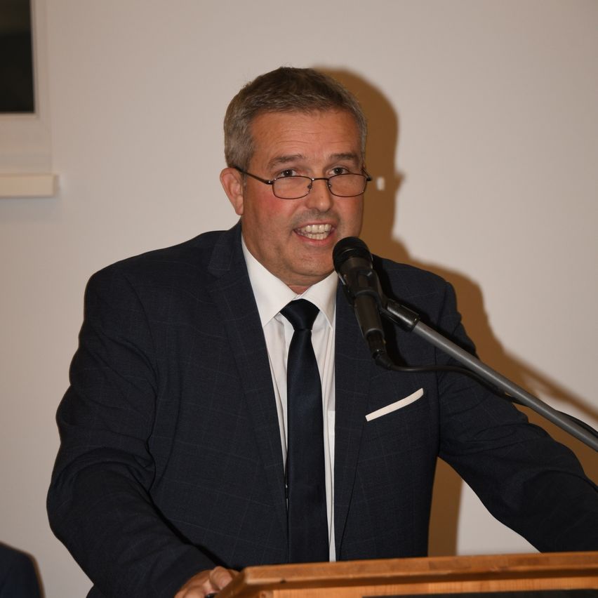 Ein Mann in einem schwarzen Anzug und einer Krawatte steht an einem Rednerpult, spricht in ein Mikrofon und hat eine weiße Wand im Hintergrund.