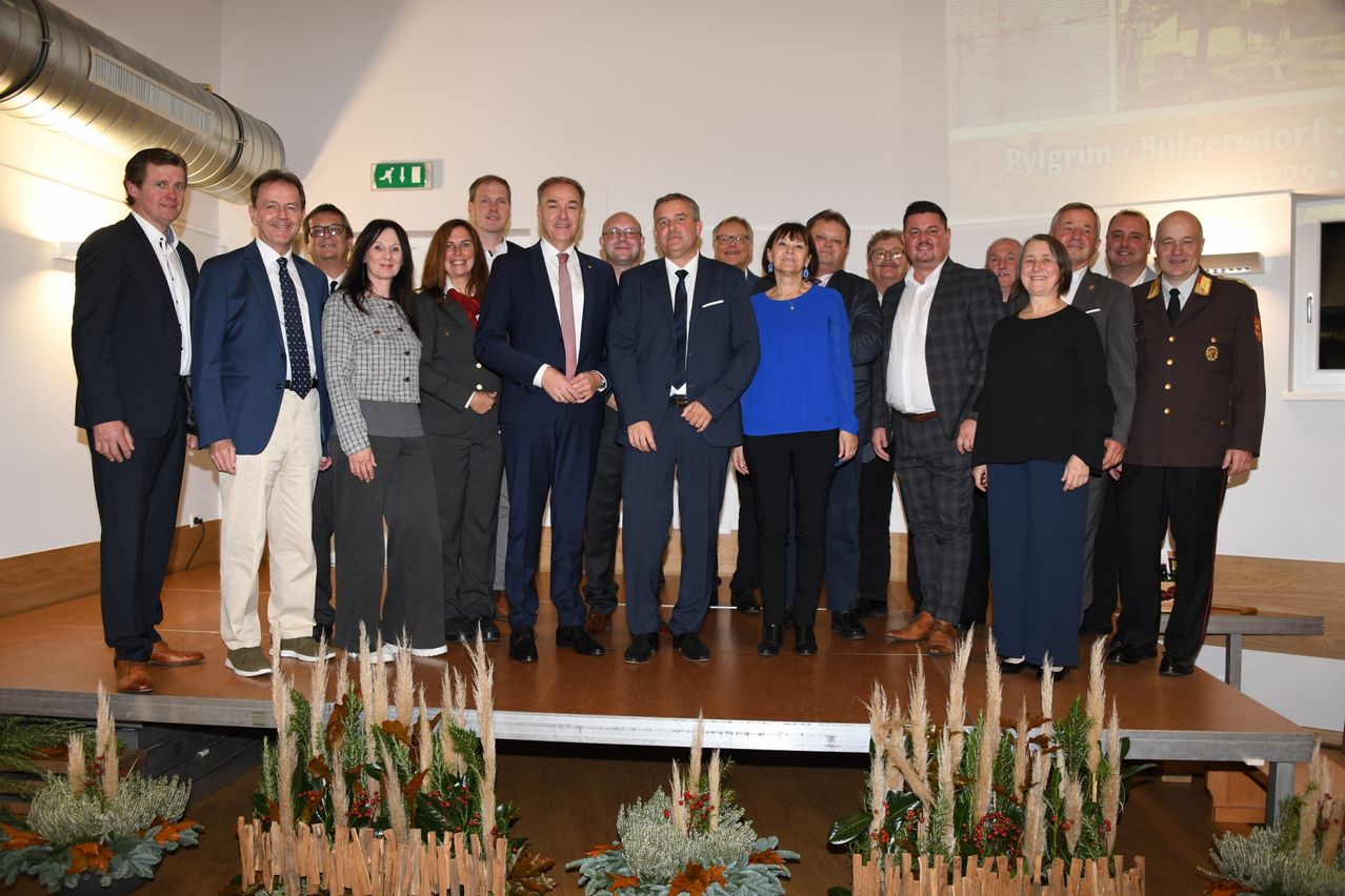 Eine Gruppe formell gekleideter Personen steht auf einer Bühne mit Pflanzen und Blumen davor. Sie scheinen für ein Foto zu posieren. Hinter ihnen zeigt eine Wand einen großen Bildschirm mit dem Wort Pilgrim.