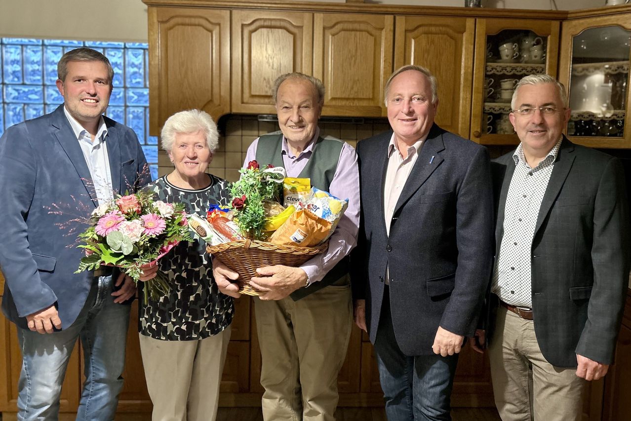 Fünf Menschen stehen in einer Küche. Ein Mann hält einen Korb mit Blumen und Essen. Eine Frau neben ihm hält einen Blumenstrauß. Sie alle lächeln.