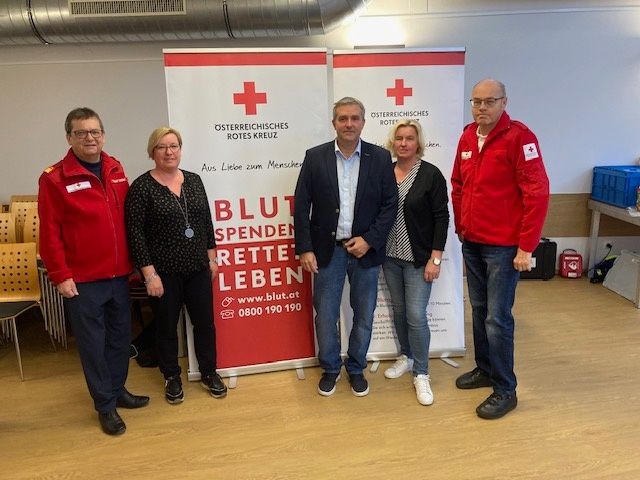 Fünf Personen stehen vor einem Banner mit der Aufschrift Blut Spenden Retten Leben und einem roten Kreuz darauf. Vier von ihnen tragen rote Jacken.