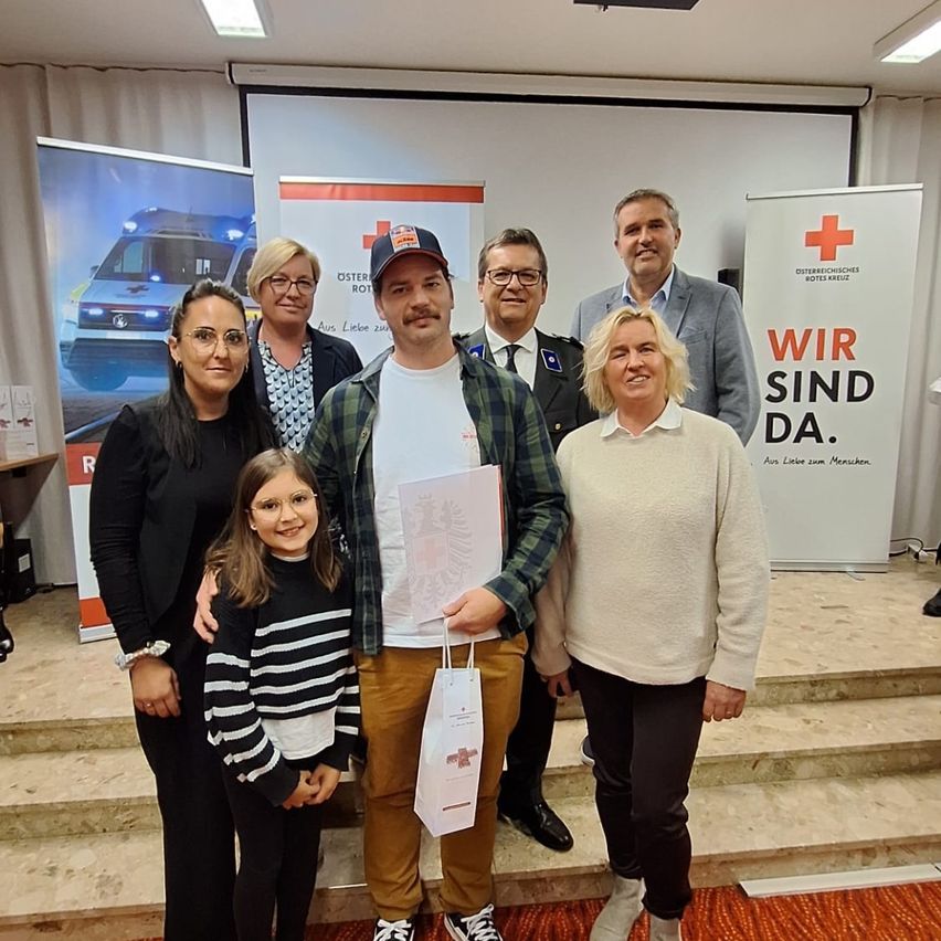 Eine Gruppe von Menschen steht auf Stufen und lächelt für ein Foto. Ein Mann in der Mitte hält einen Papierbeutel. Hinter ihnen sind Banner mit einem roten Kreuz und einem Krankenwagen zu sehen.