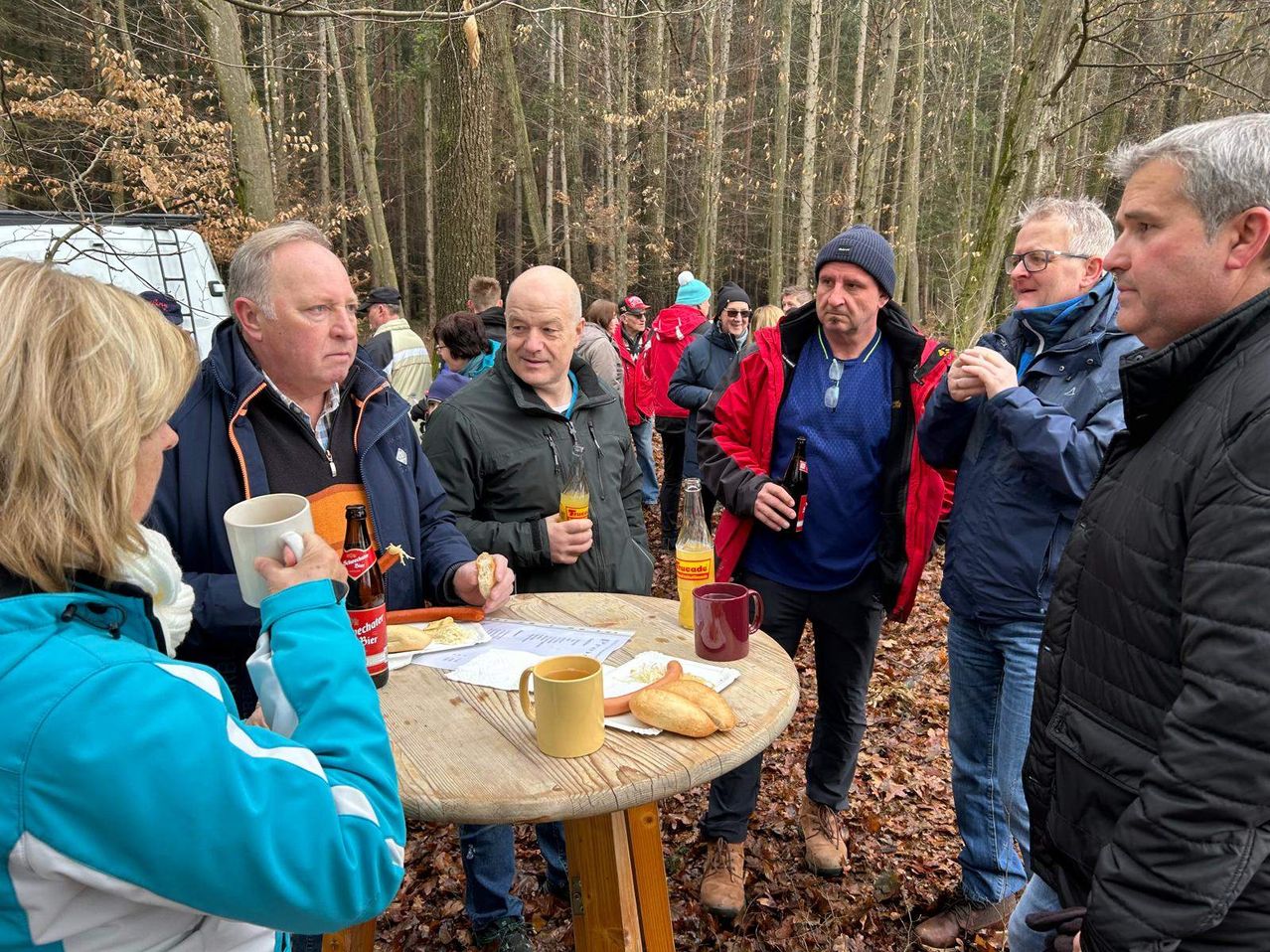 A group of people are gathered around a table in a wooded area, drinking and eating. Some hold mugs and bottles, others wear jackets and glasses. Trees surround them.