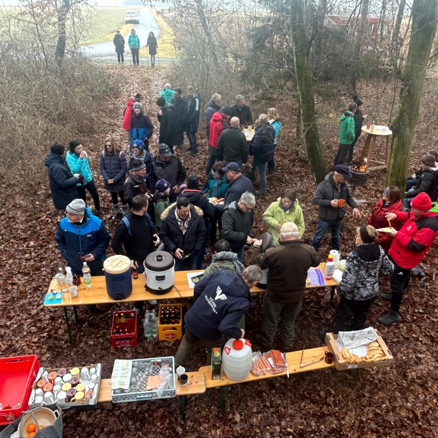 A group of people gathered in a forest for a meal, some standing around tables with food and drinks.