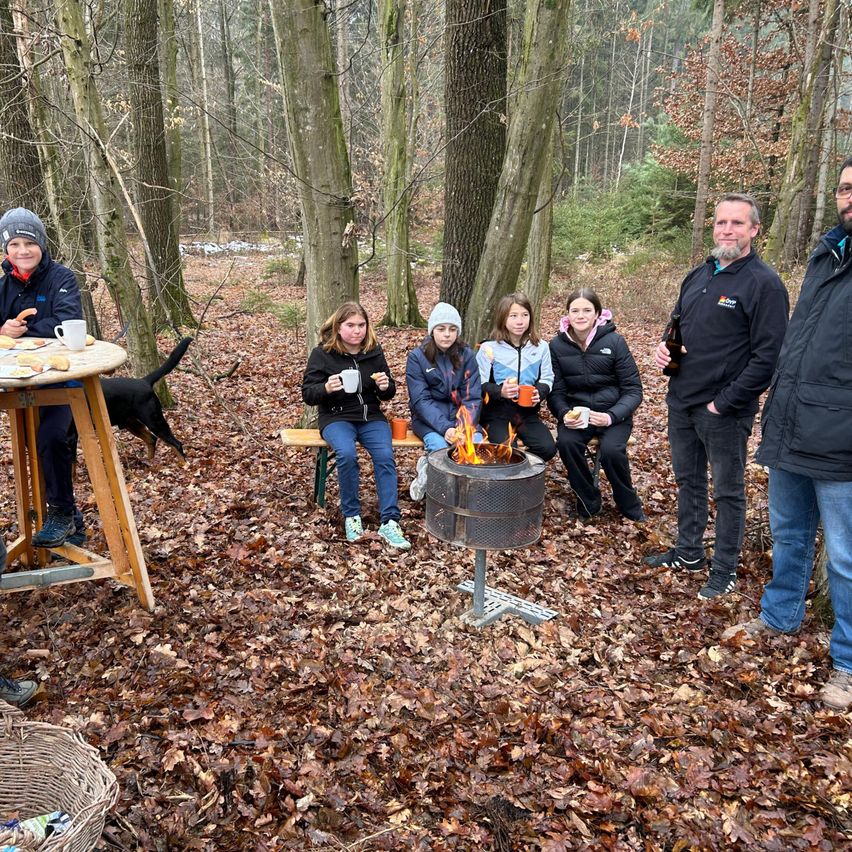 A group of people are gathered around a bonfire in a forest, some holding cups. A dog lies nearby. A table with food is on the left.