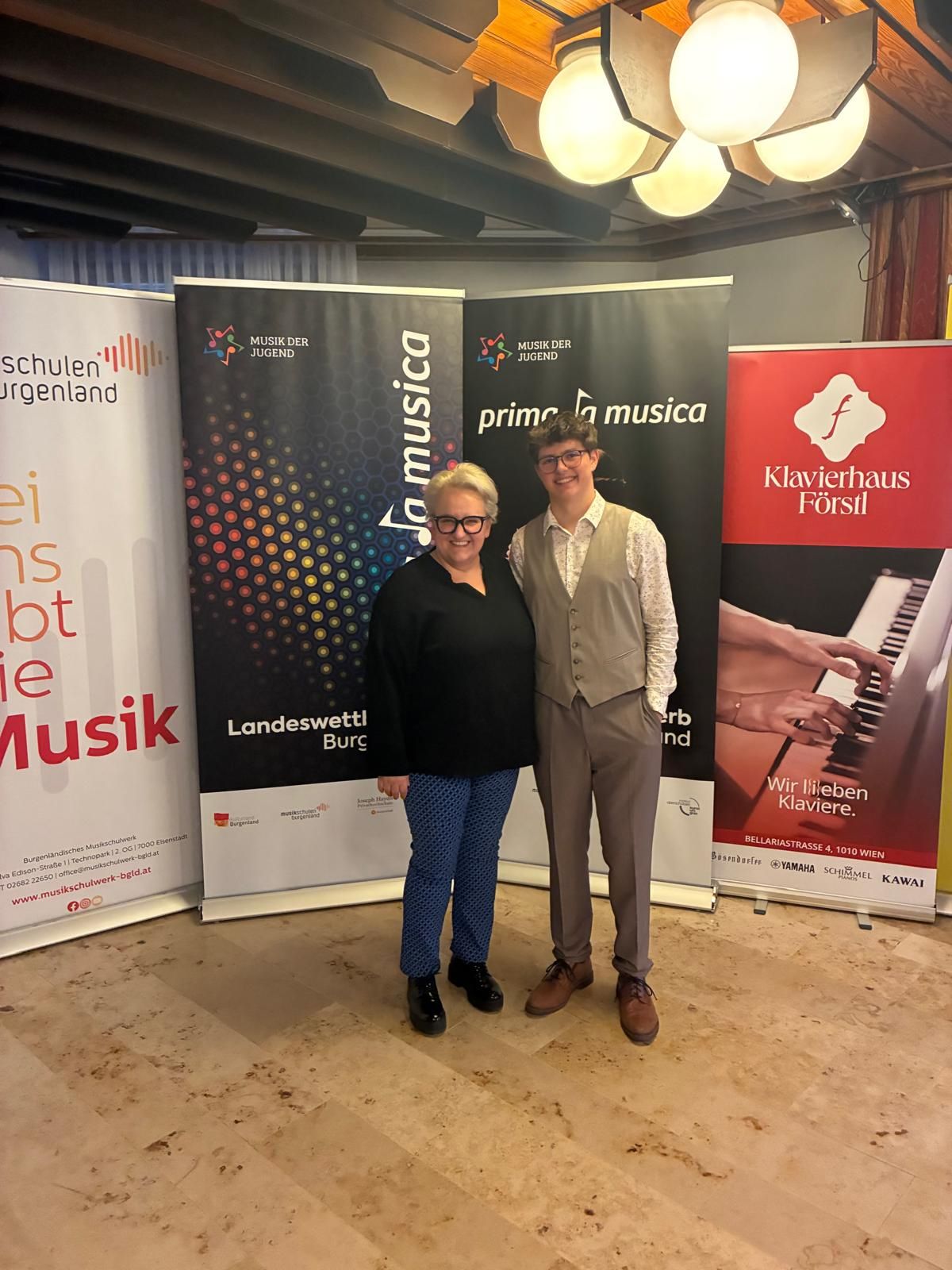 Zwei Personen stehen zusammen vor verschiedenen Bannern, eines mit einem Klavier darauf. Eine Frau trägt eine Brille und eine Weste, und ein Mann trägt eine Weste und Hose. Sie posieren für ein Foto.