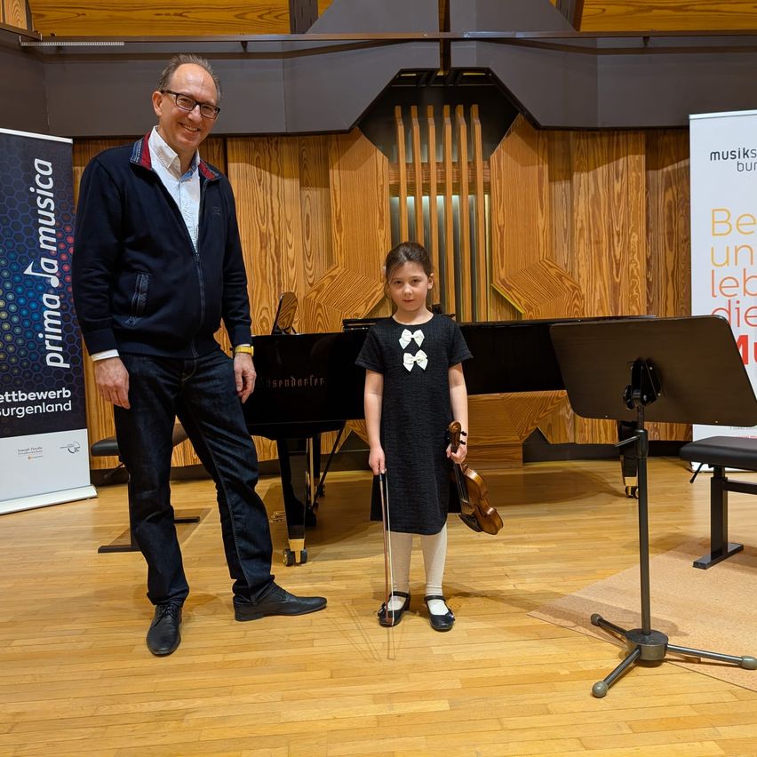 Ein Mann und ein junges Mädchen stehen neben einem Klavier mit einer Geige in einer Musikhalle mit Holzwänden.