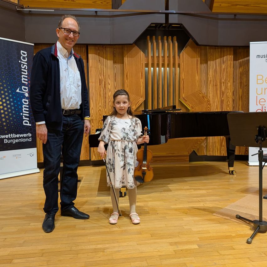 Ein Mann und ein kleines Mädchen stehen neben einem Flügel und halten eine Geige in einer Musikschule. Hinter ihnen ist eine Holzwand mit Bannern zu sehen.