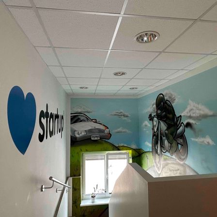 Ein Flur mit einem blauen Herz und dem Wort 'Startup' an der Wand. Die Wand hat eine Wandmalerei eines Autos und eines Motorrads. Die Decke hat quadratische Fliesen und Lampen. Es gibt ein Fenster mit einem Topf auf der Fensterbank.