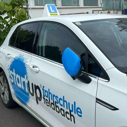Ein weißer Wagen mit 'start up fahrschule feldbach' auf der Seite ist draußen geparkt. Das Auto hat einen blauen Spiegel und ein weißes Top-Schild mit dem Buchstaben 'L'.
