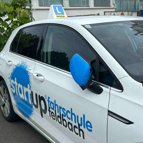Ein weißer Wagen mit 'start up fahrschule feldbach' auf der Seite ist draußen geparkt. Das Auto hat einen blauen Spiegel und ein weißes Top-Schild mit dem Buchstaben 'L'.