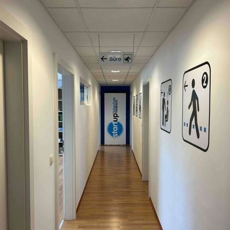 Ein Flur mit weißen Wänden, Holzböden und einem Banner mit der Aufschrift 'StartUp Feldbach'. Ein Schild, das auf das Büro zeigt, und Richtungsschilder an der rechten Wand.