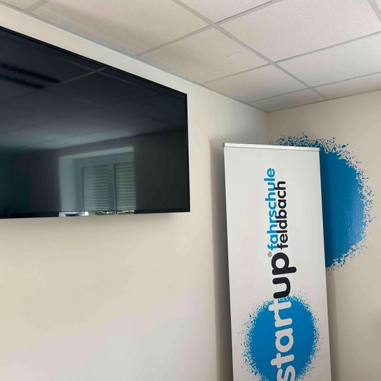 Ein Raum mit einem Flachbildfernseher an der Wand und einem Whiteboard, das 'Start up fahrschule' mit blauen Farbspritzern anzeigt.