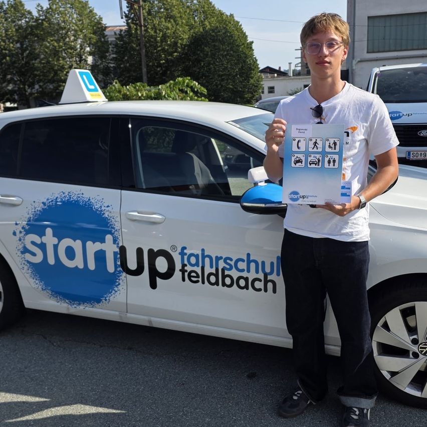A young man stands next to a white car with the text 'start up fahrschul feldbach' on the side. He holds a sign with icons and text.
