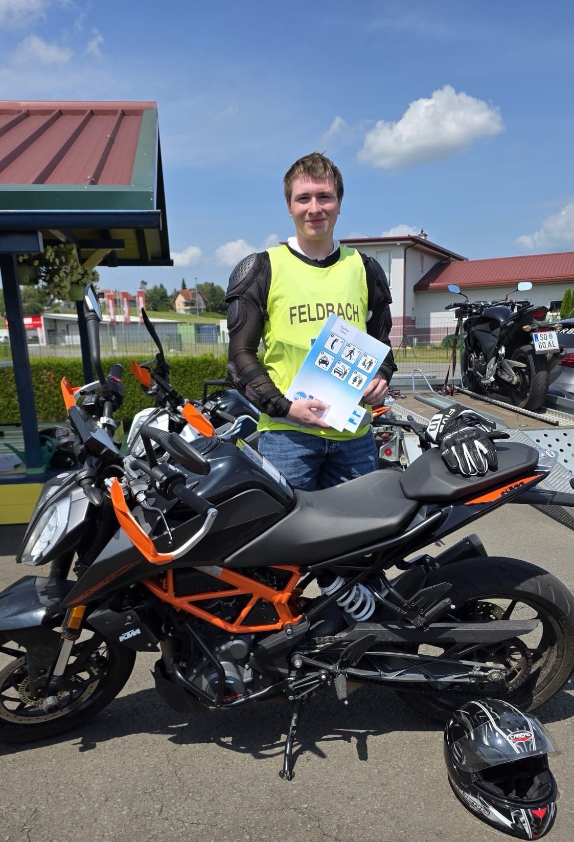 A man in a yellow vest stands next to a black and orange motorcycle, holding a brochure. Behind him, there are other motorcycles and a building with a red roof.