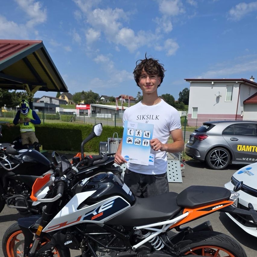 A young man stands next to a motorcycle, holding a certificate in his hands. Behind him, a building, a car, and another motorcycle are visible. The sky is blue with some clouds.