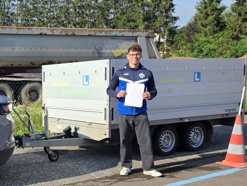 A man stands next to a trailer, holding a paper, wearing glasses and a jacket. Behind him is a large, rusty trailer.