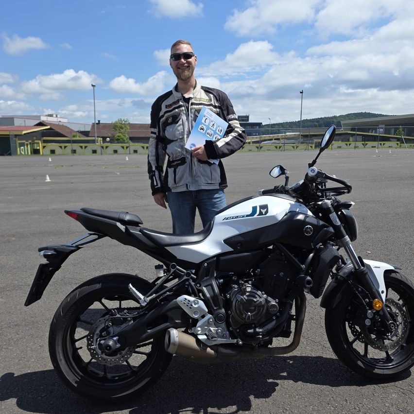 Ein Mann mit Sonnenbrille hält ein Büchlein in der Hand, während er neben einem Motorrad auf einem Parkplatz steht. Hinter ihm befindet sich ein Gebäude und Berge.