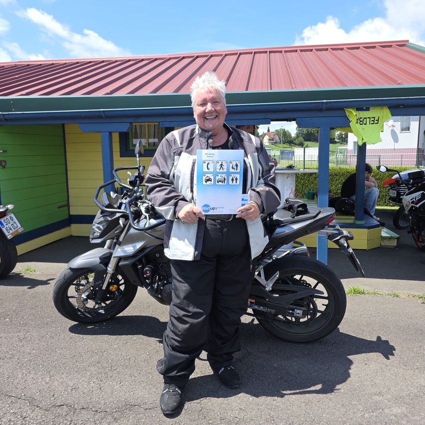 Ein Mann steht vor einem Motorrad und hält ein blaues Dokument mit Symbolen in der Hand und lächelt. Dahinter befindet sich ein Gebäude mit rotem Dach und gelber Wand.