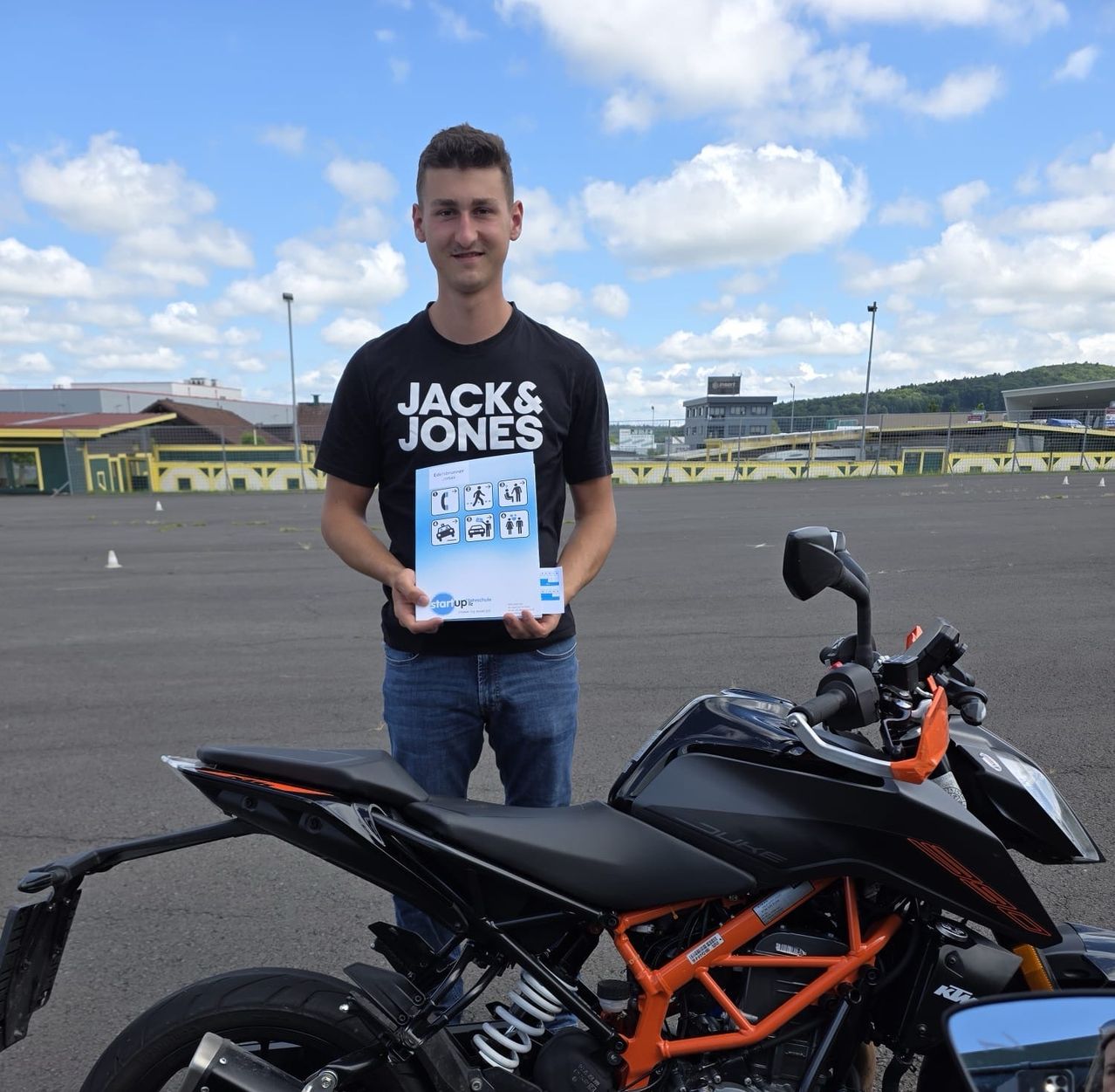 Ein Mann steht neben einem schwarzen Motorrad auf einem Parkplatz. Er hält ein Buch in beiden Händen mit dem Text Jack & Jones.