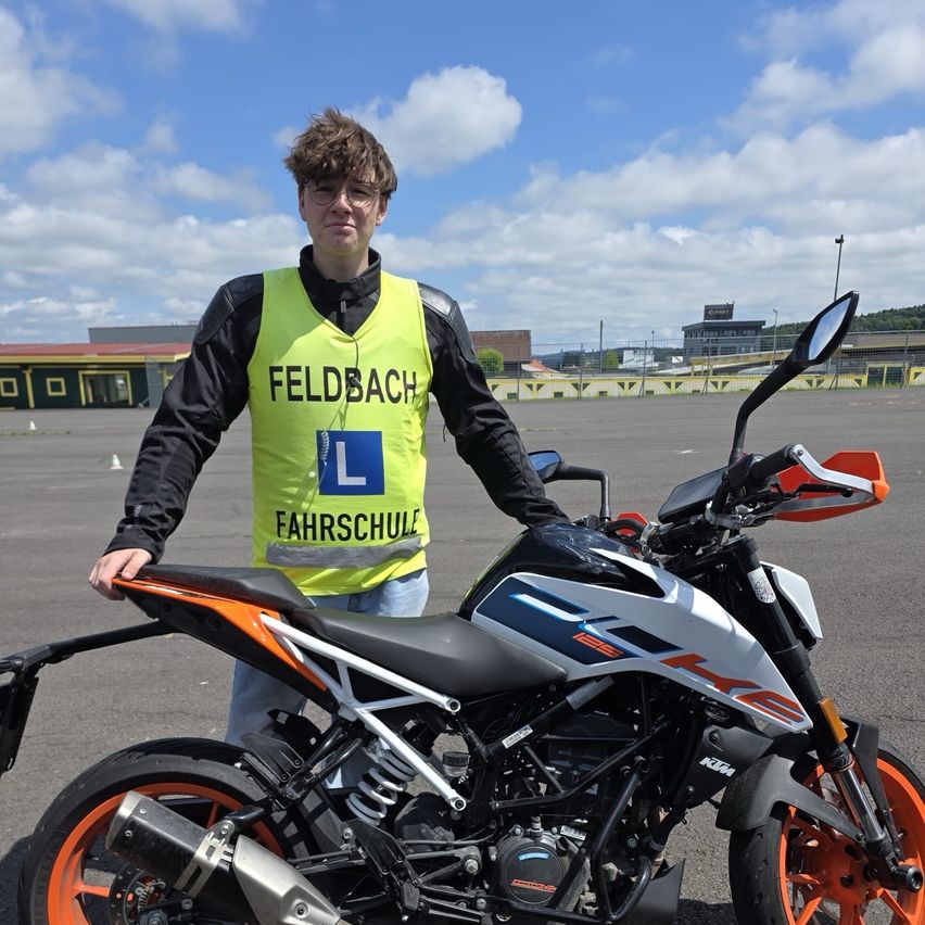 Ein Mann in einer Warnweste mit den Buchstaben L steht neben einem Motorrad. Das Motorrad ist orange und schwarz.