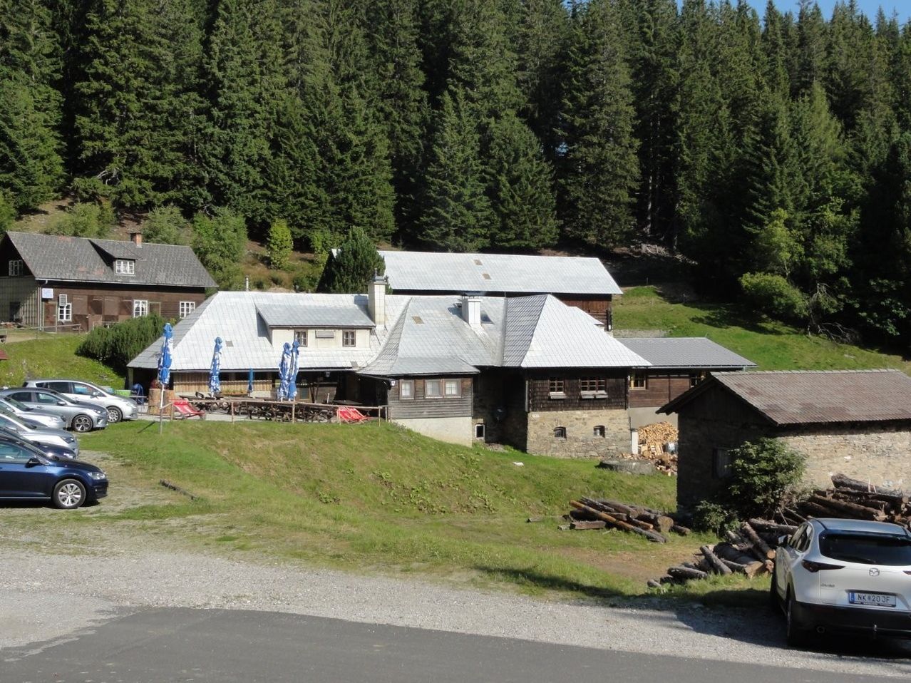 Ein Berghaus mit grauem Dach und Schornsteinen liegt zwischen Bäumen. Im Freien sind Tische mit blauen Sonnenschirmen aufgestellt.