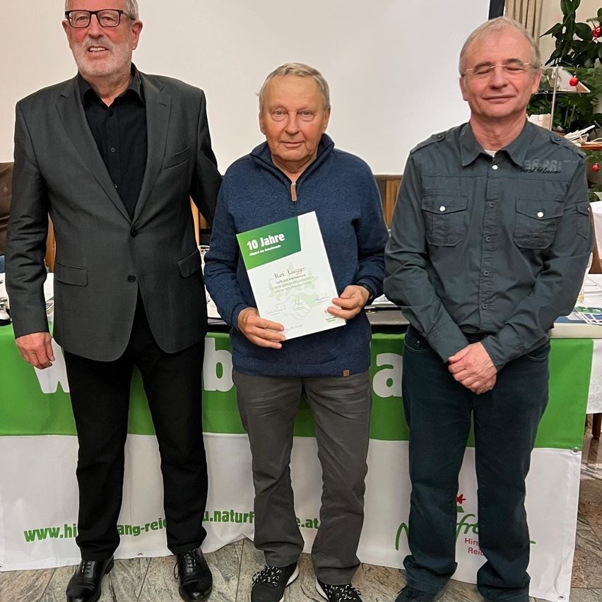 Drei Männer stehen hinter einem Tisch mit einem Banner. Der Mann in der Mitte hält ein Zertifikat mit der Aufschrift '10 Jahre'. Der Mann links trägt einen Anzug, während die anderen beiden Freizeithemden tragen.