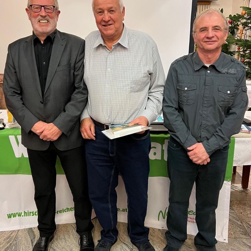 Drei Männer stehen vor einem Hintergrund mit einem Website-Logo und lächeln. Der Mann in der Mitte hält ein Buch. Dahinter ist ein Weihnachtsbaum zu sehen.