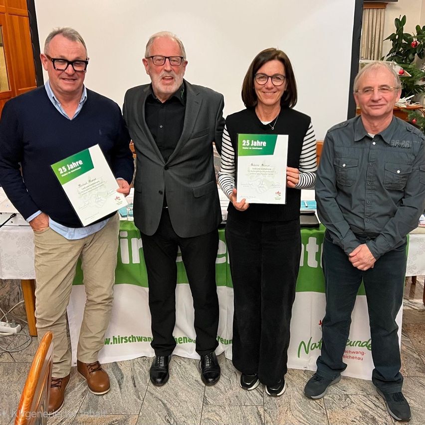 Vier Personen stehen vor einem Tisch, lächeln und halten Urkunden in den Händen. Der Tisch ist mit einer grün-weißen Tischdecke bedeckt. Die Frau in der Mitte hält eine Urkunde mit '25 Jahre' darauf. Der Mann links trägt eine Brille und einen braunen Schuh.