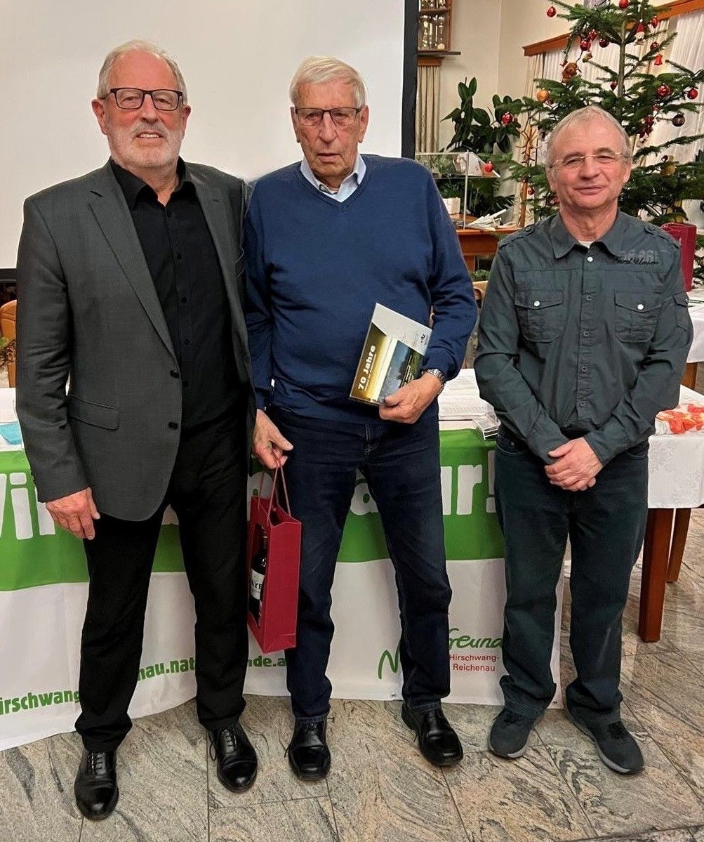 Drei Männer stehen nebeneinander vor einem Tisch mit einer weißen Tischdecke. Der Mann auf der linken Seite hält eine rote Tasche mit einer Flasche darin. Der Mann in der Mitte hält ein Buch. Der Mann auf der rechten Seite trägt ein grünes Hemd. Dahinter steht ein Weihnachtsbaum.