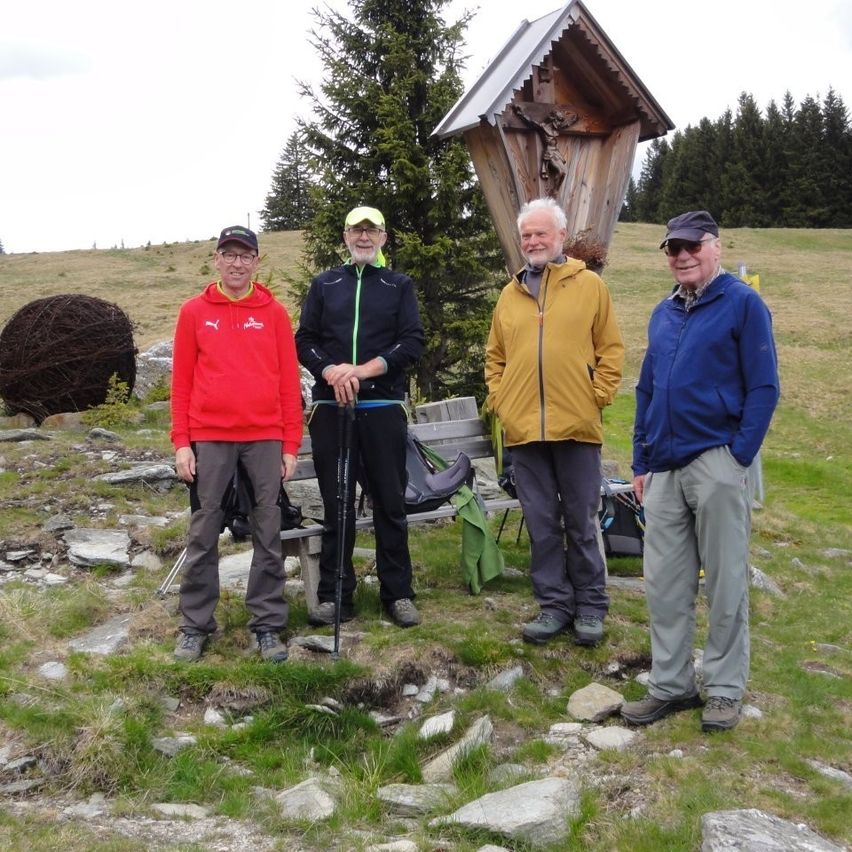 Vier ältere Männer stehen vor einer hölzernen Kapelle in einem grasigen Bereich. Jeder Mann trägt eine Brille und hält einen Wanderstock. Hinter ihnen steht eine Bank mit Rucksäcken. Bäume und eine grasige Anhöhe sind im Hintergrund.