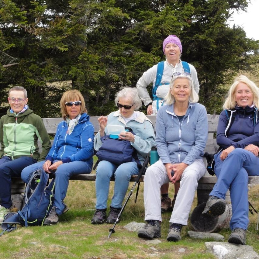 Fünf Frauen und ein Mann sitzen auf einer Bank im Wald, alle tragen Sonnenbrillen und Jacken. Hinter ihnen stehen hohe Bäume.