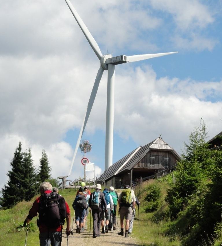 Eine Gruppe von Wanderern mit Rucksäcken und Hüten geht auf einem Grashügel, mit einer Windturbine und einem Haus im Hintergrund.