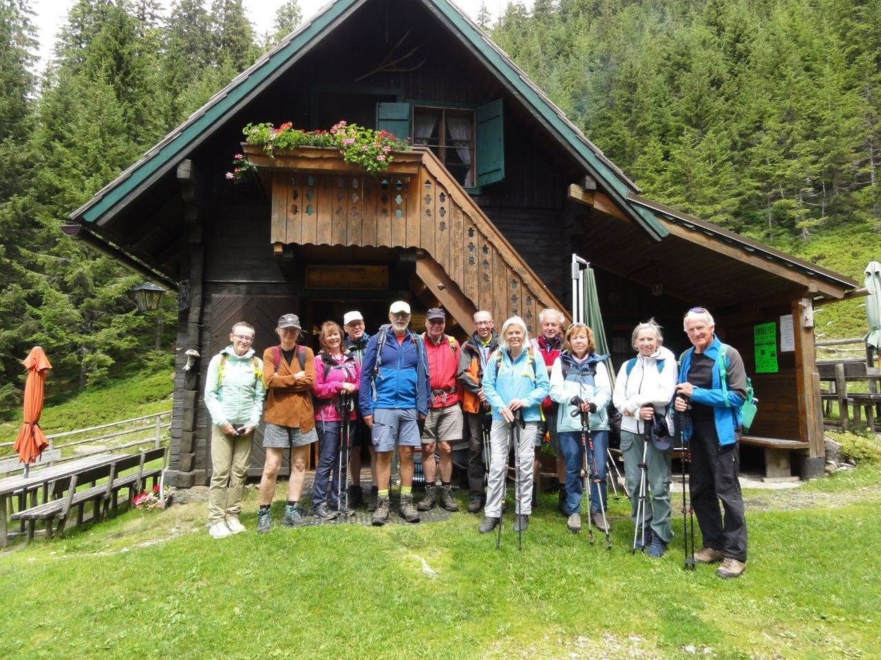 Eine Gruppe von Wanderern in Outdoor-Kleidung, die vor einem Holzchalet mit Balkon und Blumen an einem sonnigen Tag stehen.