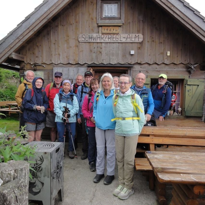 Eine Gruppe von Wanderern in Outdoor-Kleidung steht vor einem Holzgebäude mit dem Schild 'Schwarzriegl-Alm'. Sie lächeln und scheinen für ein Foto zu posieren.