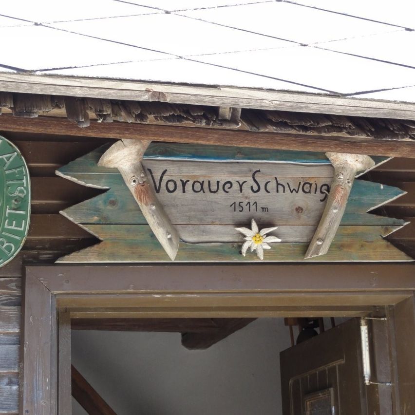 Ein hölzernes Schild mit dem Text 'Vorauer Schwaig' und einer weißen Blume ist über dem Eingang eines Holzgebäudes angebracht. Das Schild ist mit hölzernen Geweihen am Dach befestigt.