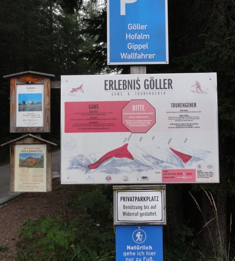 Ein Schild mit einer Karte des Gebiets, das 'Erlebenis Goller' anzeigt, und ein rotes Stoppschild mit dem Wort 'Bitte'. Links befinden sich zwei kleine Holzschilder, eines mit einem Bild eines Hauses und das andere mit einem Kalender.