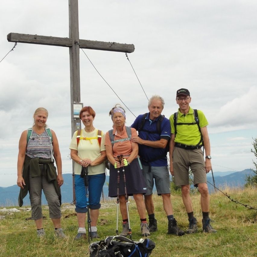 Fünf Wanderer mit Rucksäcken und Stöcken stehen am Gipfelkreuz vor bewölktem Himmel.