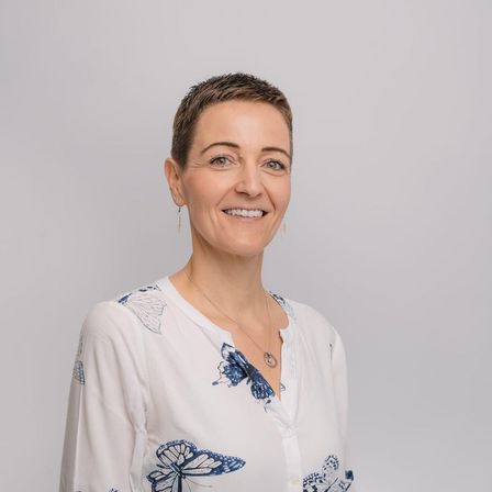 Eine Frau mit kurzen Haaren lächelt in die Kamera. Sie trägt ein weißes Hemd mit blauen Schmetterlingsaufdrucken und eine Halskette. Der Hintergrund ist schlicht und weiß.