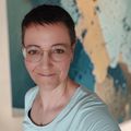 Eine Frau mit kurzen Haaren und Brille lächelt vor einem blau-gelben abstrakten Gemälde.